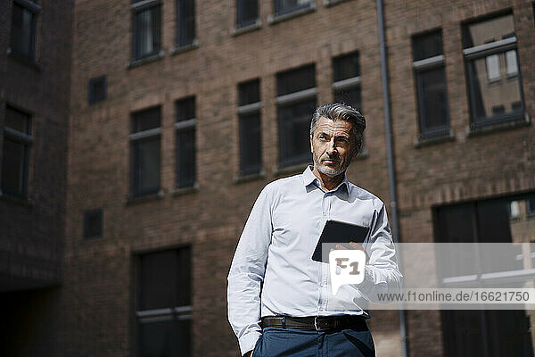 Businessman looking away while using digital tablet against building