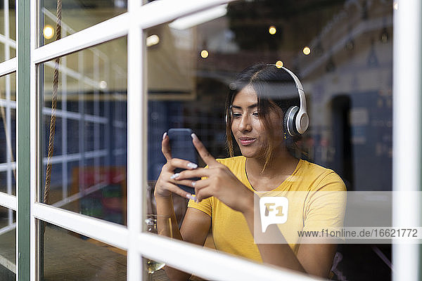 Schöne junge Frau hört Musik und benutzt ihr Smartphone in einer Bar  gesehen durch ein Fenster