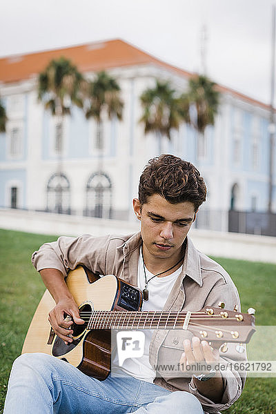 Handsome young man practicing guitar while sitting on grass at park