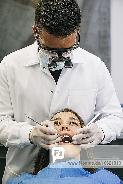 Männlicher Zahnarzt mit chirurgischer Lupenbrille bei der Untersuchung der Zähne einer jungen Patientin in der Klinik