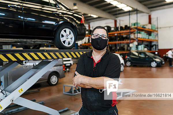 Male mechanic wearing mask with arms crossed standing in workshop