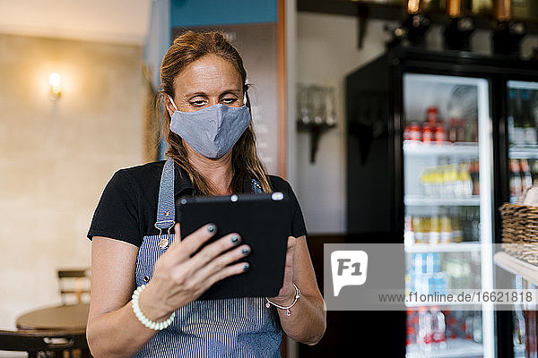Weibliche Besitzerin mit Gesichtsschutzmaske bei der Nutzung eines digitalen Tablets in einem Café