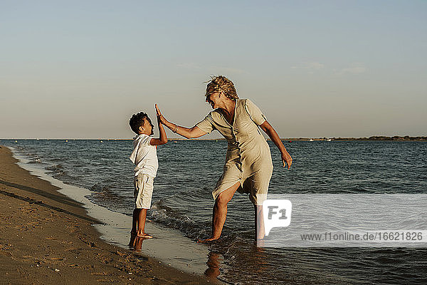 Glückliche Großmutter und Enkel machen High Five zueinander am Strand während des Sonnenuntergangs