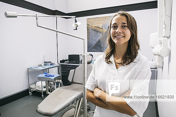 Lächelnde junge Zahnarzthelferin steht mit verschränkten Armen in einer Klinik