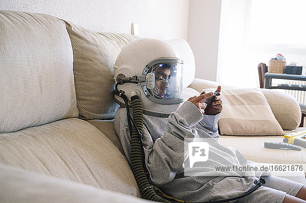 Junge mit Weltraumhelm  der ein Handheld-Videospiel spielt  während er sich auf dem Sofa zu Hause entspannt