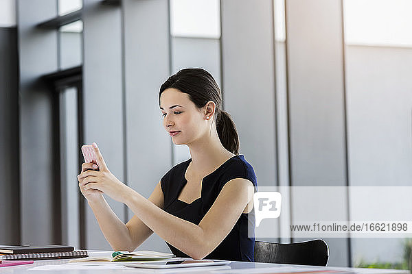 Selbstbewusste Geschäftsfrau  die ein Smartphone benutzt  während sie im Büro am Schreibtisch sitzt