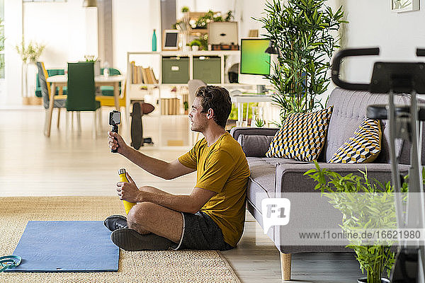 Fitnesstrainer  der ein Stativ mit Mobiltelefon hält  während er zu Hause sitzt