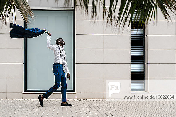 Glücklicher männlicher Unternehmer  der einen blauen Anzug hält  während er in der Stadt auf dem Bürgersteig geht