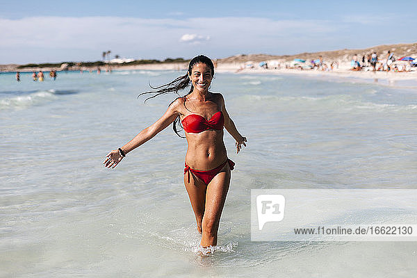 Cheerful mid adult woman wearing bikini walking in sea against sky