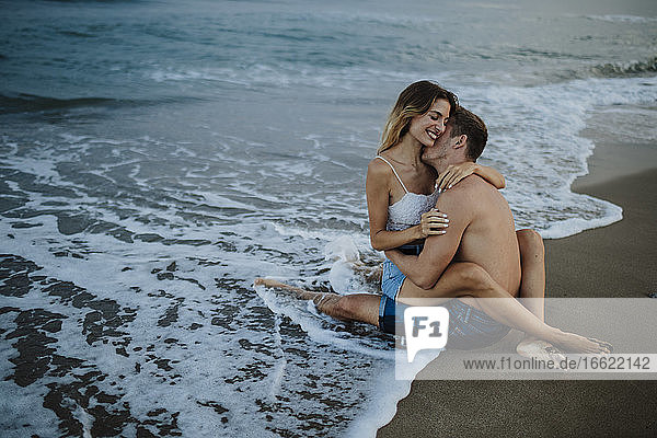 Paar umarmt sich  während es am Strand am Rande des Wassers sitzt
