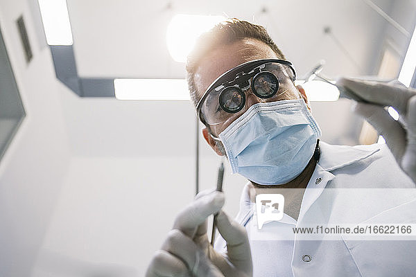 Männlicher Zahnarzt in chirurgischer Lupenbrille und Maske mit medizinischen Instrumenten während der Behandlung in der Klinik