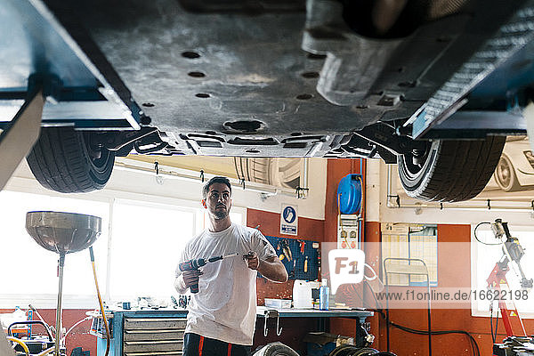 Young mechanic holding work tool looking at car while standing in auto repair shop