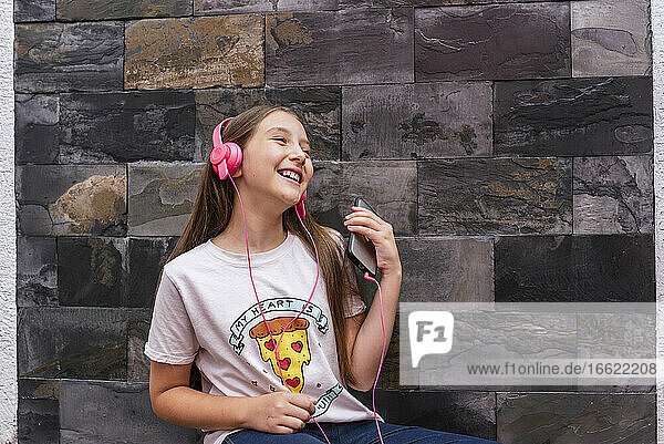 Fröhliches Mädchen mit Kopfhörer hört Musik  während sie zu Hause an der Wand sitzt