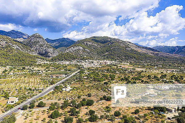 Luftaufnahme einer Straße in Richtung eines Dorfes in der Nähe einer Gebirgskette bei bewölktem Himmel  Caimari  Mallorca  Spanien