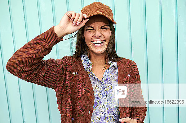 Glückliche junge Frau  die eine Mütze hält und vor einer blauen Metalltür steht