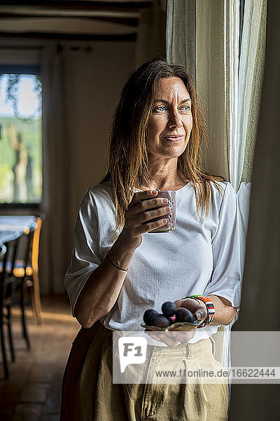 Thoughtful female nutritionist holding drink and fruits while looking through window