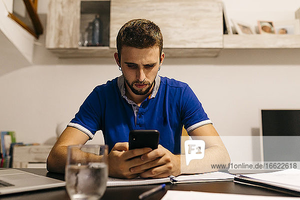 Gut aussehender junger Mann  der ein Smartphone benutzt  während er mit einem Arbeitsbuch am Tisch sitzt