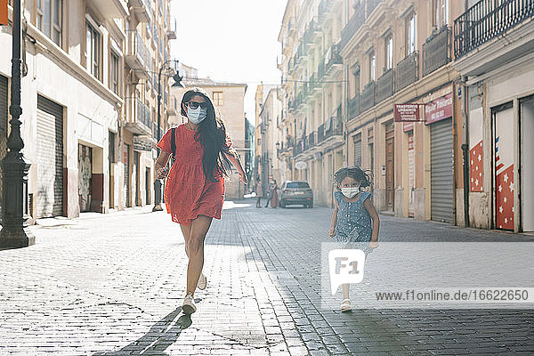 Fröhliche Mutter und Tochter mit Masken laufen auf der Straße in der Stadt