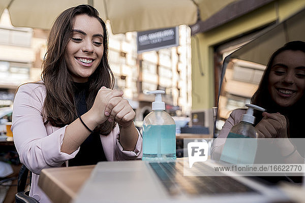 Smiling young businesswoman using hand sanitizer in cafe during COVID-19 outbreak