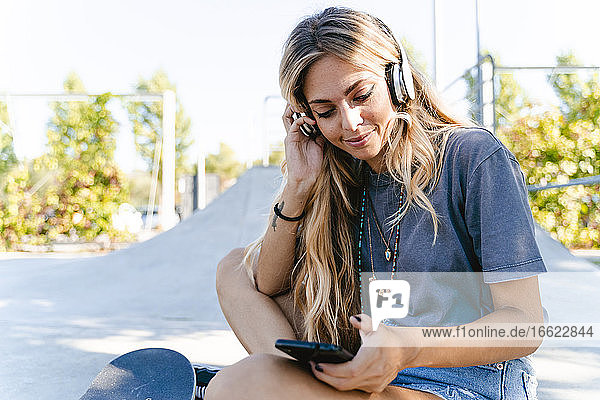 Junge blonde Frau hört Musik über Kopfhörer  während sie ihr Smartphone im Skateboard-Park benutzt