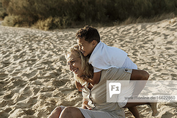 Enkel  der seine Großmutter von hinten umarmt  während er im Sand am Strand sitzt