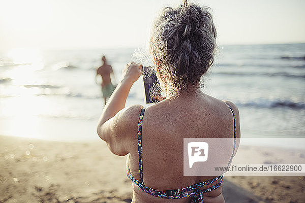 Frau  die am Strand stehend ein Foto mit ihrem Mobiltelefon macht