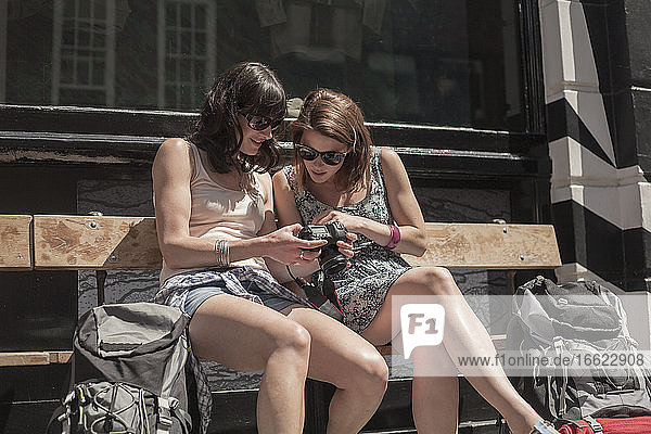 Curious woman looking at camera held by female friend while sitting on bench in city