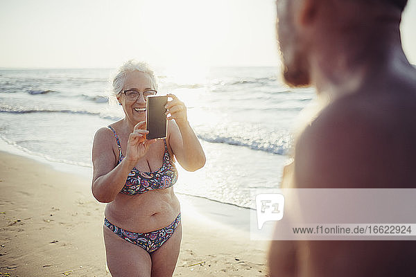 Ältere Frau im Bikini fotografiert einen Mann am Strand