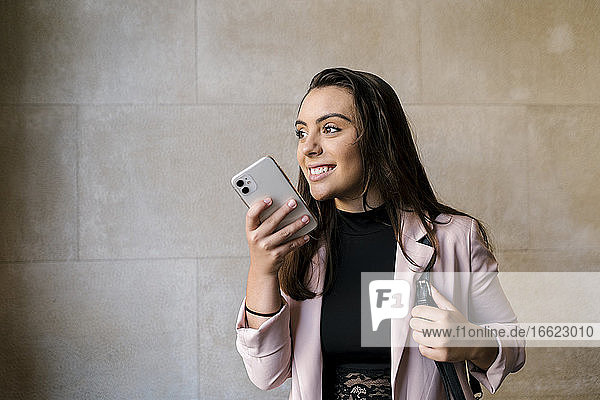 Smiling young businesswoman talking through smart phone against wall in cafe