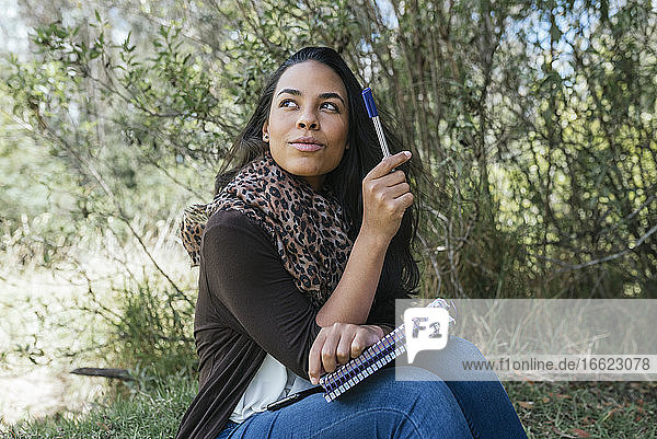 Nachdenkliche junge Frau  die mit Tagebuch und Stift im Park sitzt und wegschaut