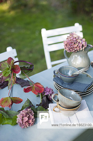 Geschirr und DIY-Lampenschirm aus Blättern und Hortensienblüten auf einem Couchtisch im Garten liegend