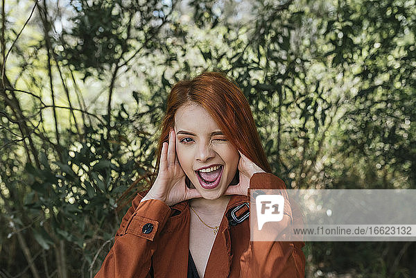 Playful young redhead woman winking while standing at park