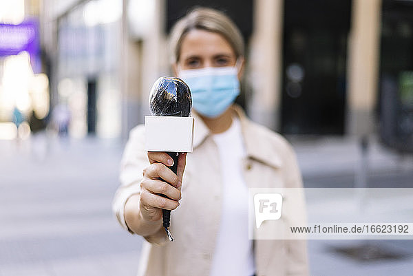 Weibliche Journalistin mit Maske  die ein Mikrofon hält  während sie auf einer Straße in der Stadt steht
