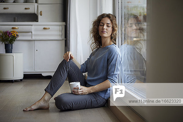 Mid adult woman with coffee cup sitting while leaning on glass door at home