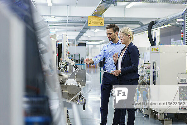 Confident young engineer discussing with businesswoman while pointing at machinery in factory