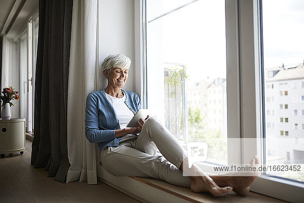 Glückliche ältere Frau  die ein digitales Tablet benutzt  während sie zu Hause am Fenster sitzt