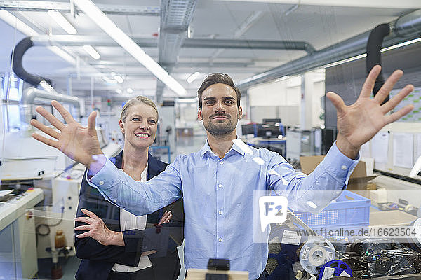 Confident young businessman gesturing at glass interface to smiling businesswoman in factory