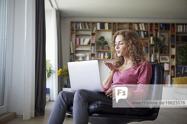 Frau bläst Kuss zu Videoanruf auf Laptop  während sie zu Hause sitzt