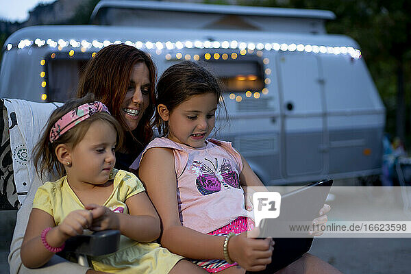 Lächelnde Mutter mit Töchtern  die ein digitales Tablet benutzen  während sie in der Abenddämmerung an einem Wohnmobil sitzen