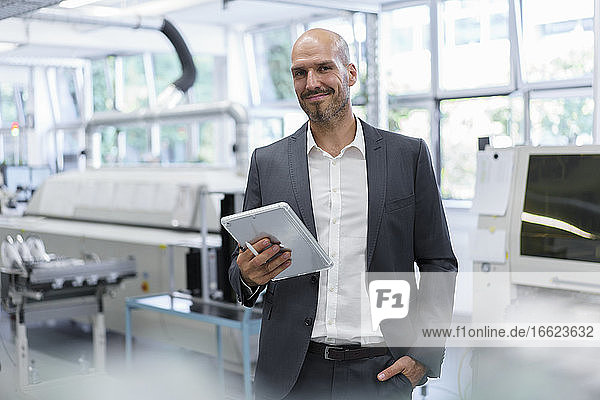Smiling confident businessman holding digital tablet while standing with hand in pocket at bright factory
