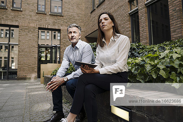 Ein Kollege hält ein digitales Tablet in der Hand  während er auf einer Stützmauer vor einem Gebäude sitzt