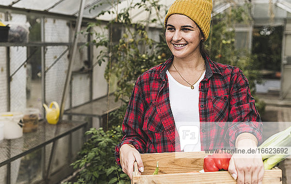 Smiling woman carrying crate with vegetables looking away while standing against greenhouse