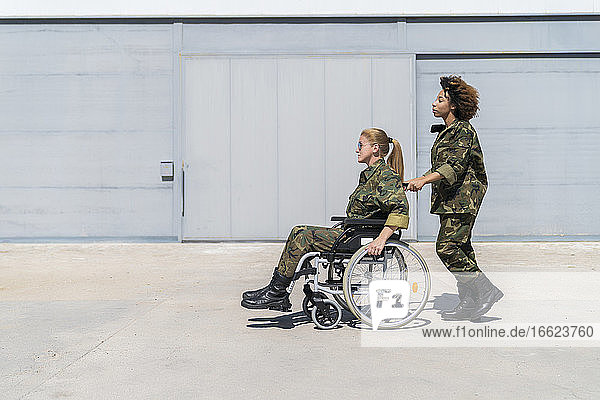 Junge Soldatin schiebt ihren Kollegen im Rollstuhl auf einem Militärstützpunkt an einem sonnigen Tag