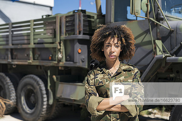 Selbstbewusste junge Soldatin  die an einem sonnigen Tag mit verschränkten Armen vor einem Lastwagen auf einem Stützpunkt steht