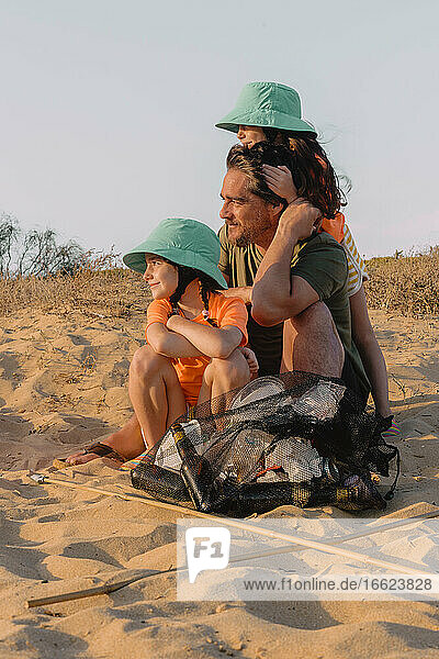 Vater und Töchter sitzen auf Sand mit Müllsack