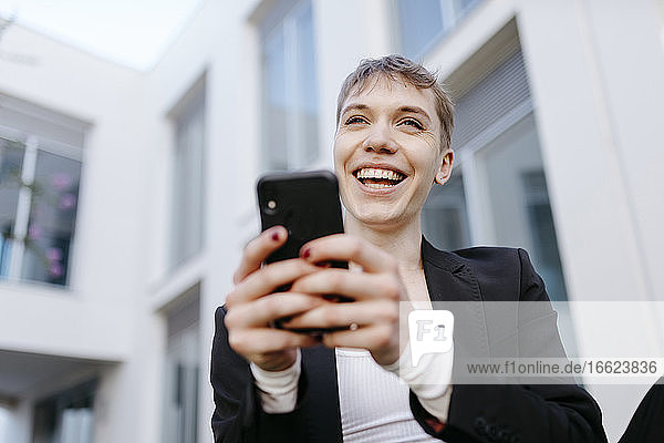 Nahaufnahme eines fröhlichen trans jungen Mannes  der ein Mobiltelefon benutzt  während er an einem Gebäude sitzt