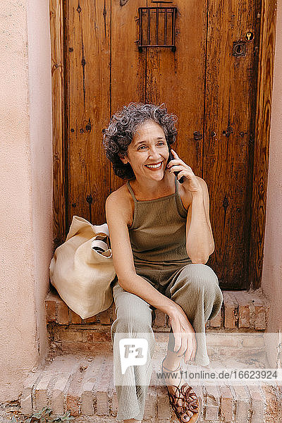 Mature woman talking on smart phone while sitting on doorway