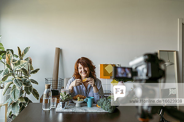 Lächelnde Frau zeigt frisches Obst  während sie zu Hause einen Vlog mit der Kamera macht