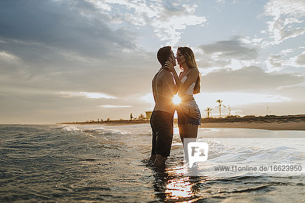 Mann und Frau küssen sich  während sie bei Sonnenuntergang am Strand im Wasser stehen