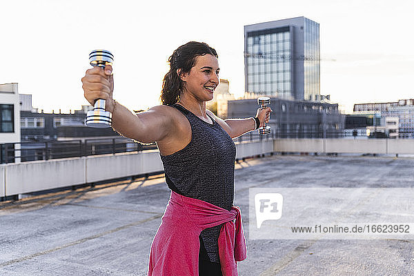 Lächelnde junge Frau mit ausgestreckten Armen hebt Hanteln auf einer Terrasse bei Sonnenuntergang